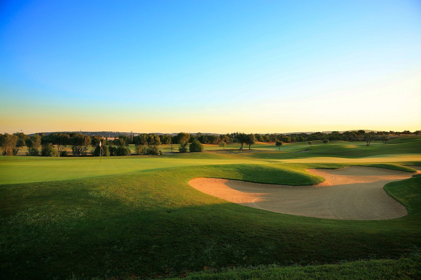 Golf course fairway with sand bunker at sunset, tree line in background under clear blue sky