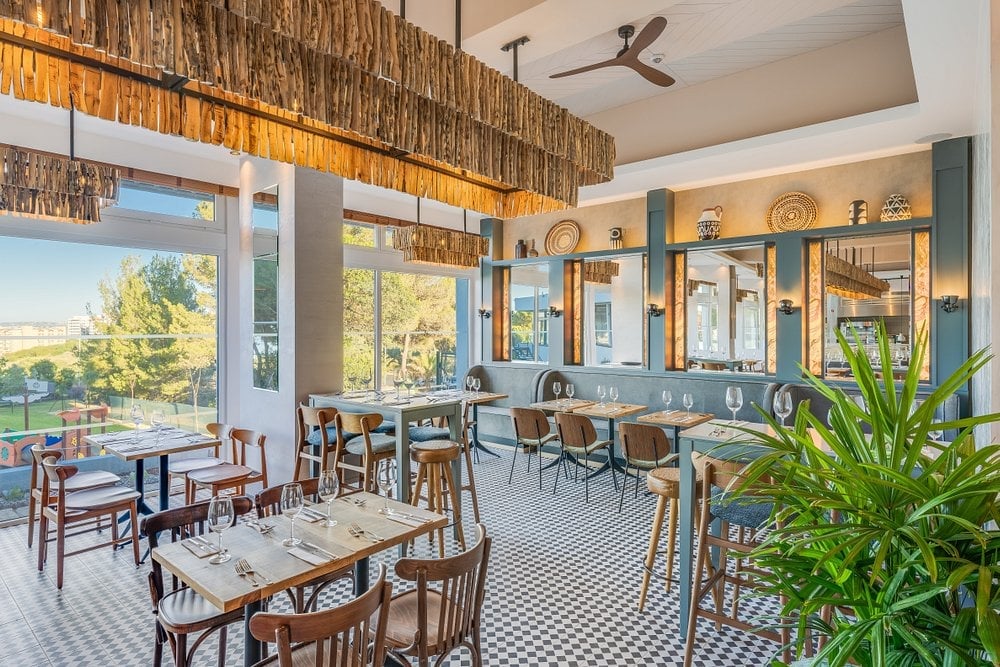 Bright restaurant interior with wooden tables, woven ceiling details, blue-green accents, checkered flooring, and large windows overlooking trees.