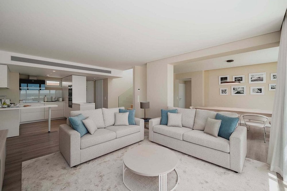 Modern open-concept living room with two cream-colored sofas, blue accent pillows, white oval coffee table, and adjacent kitchen area with dark wood flooring.