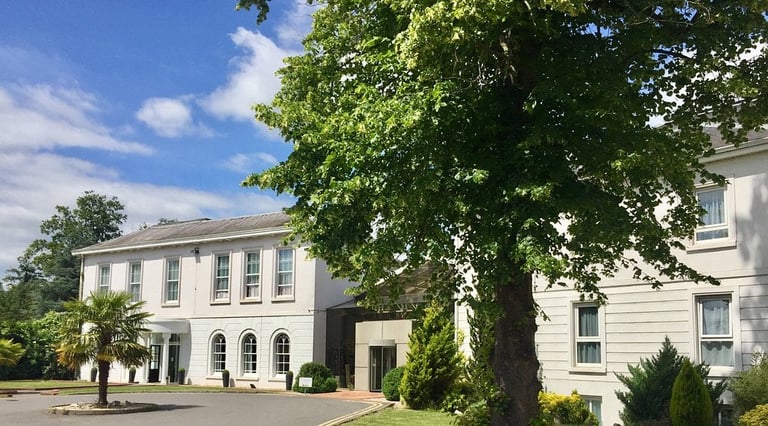 Two-story white mansion with columns and large mature trees in front, sunny day with blue sky