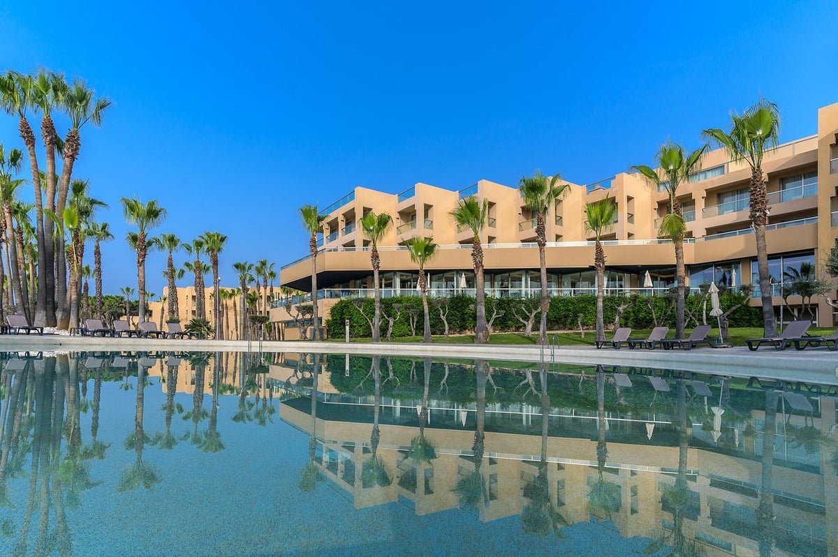 Modern terracotta-colored resort building reflected in a turquoise pool with palm trees lining the property under blue sky