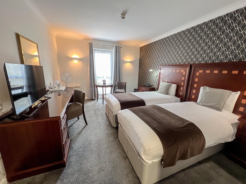 Modern hotel room with two queen beds, dark wood headboards, desk area with TV, and patterned wallpaper
