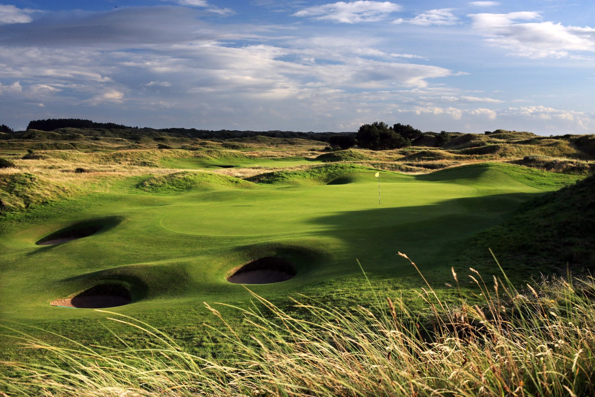 A scenic links-style golf course with rolling green fairways, multiple deep sand bunkers, and rough coastal grassland under a partly cloudy sky.