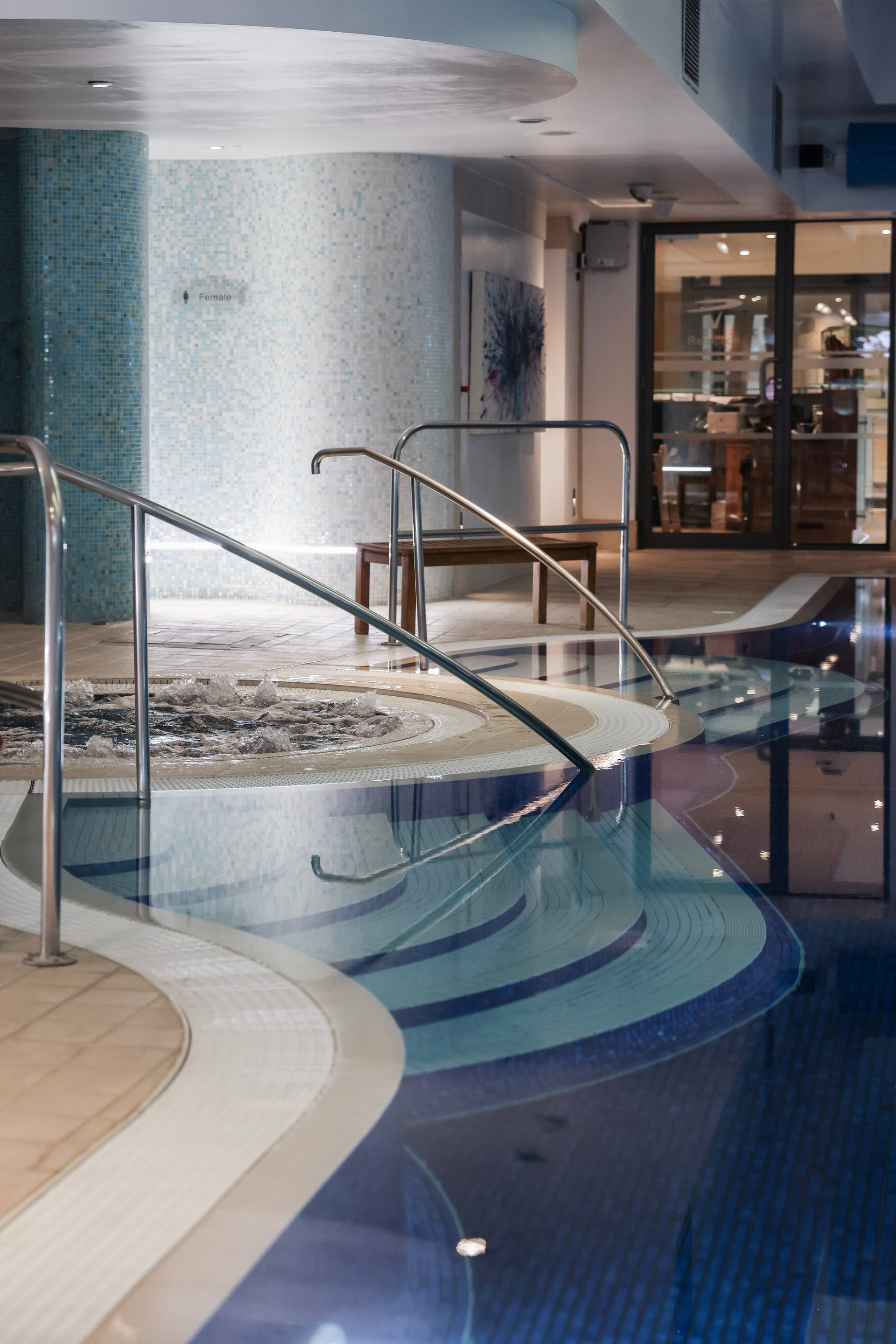 Modern indoor spa with curved swimming pool, metal railings, and glass doors opening to a wellness lounge area