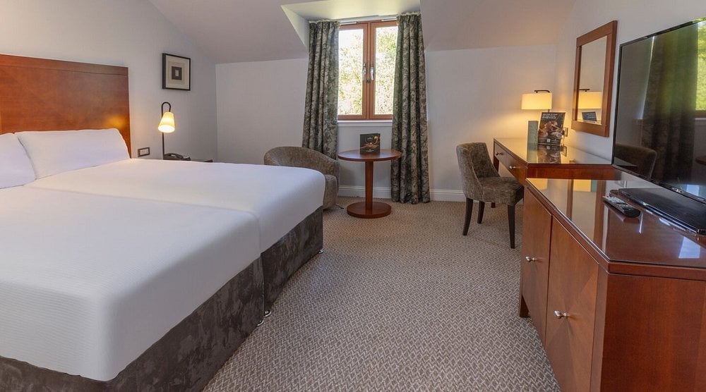 Hotel bedroom with queen bed, wooden furniture, TV, desk, and window with curtains