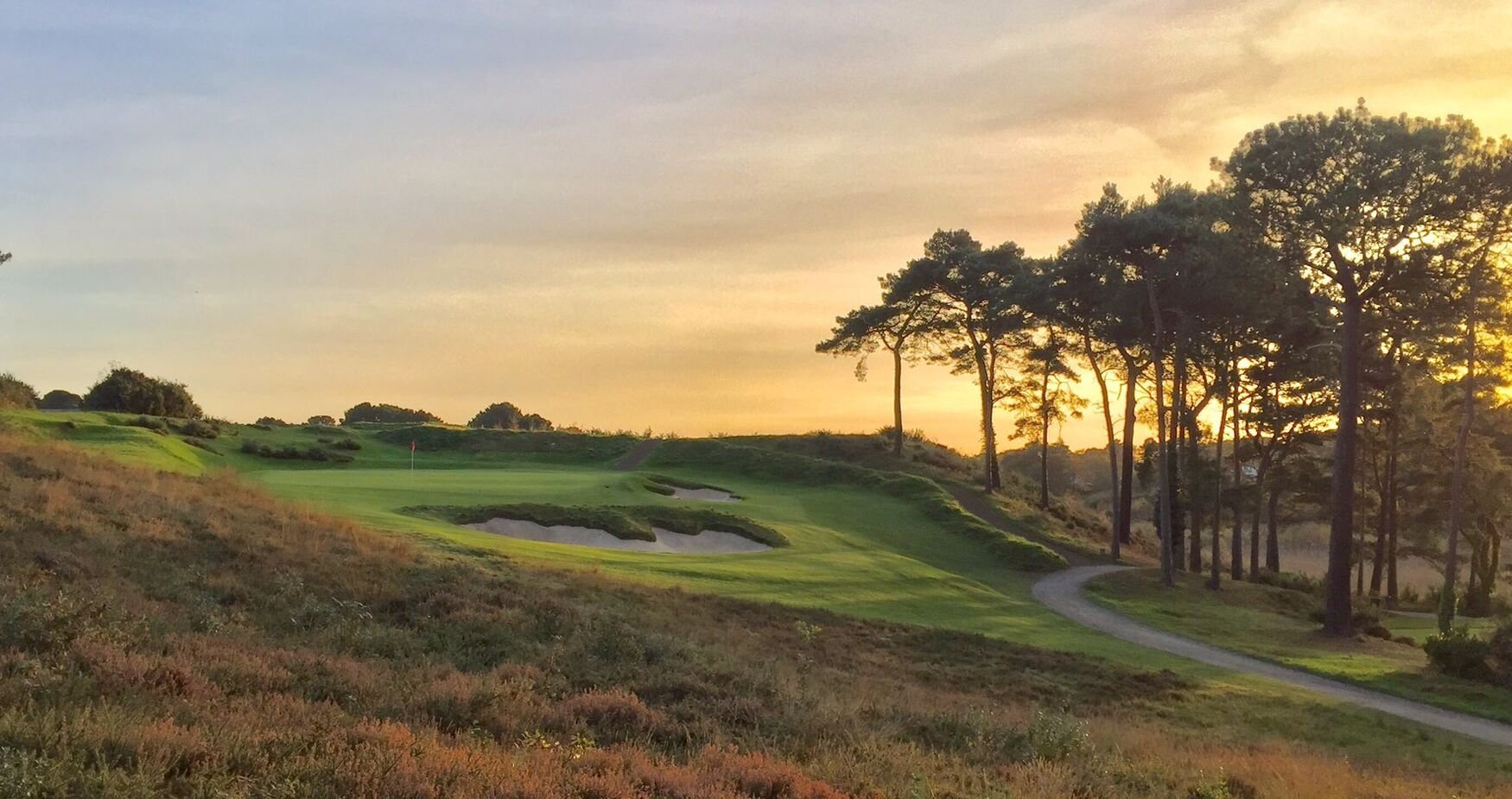 Golf course fairway at sunset with tree-lined edges and golden sky over rolling green landscape