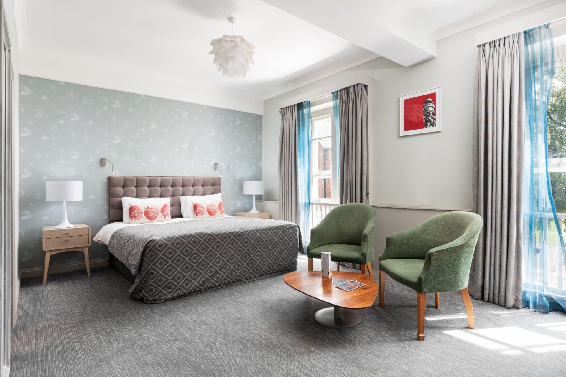 Modern bedroom with upholstered bed, green armchair, side tables with lamps, and large windows with curtains