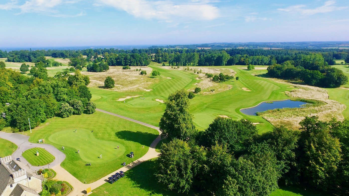Aerial view of a lush green golf course with manicured fairways, scattered trees, and a small pond under blue sky