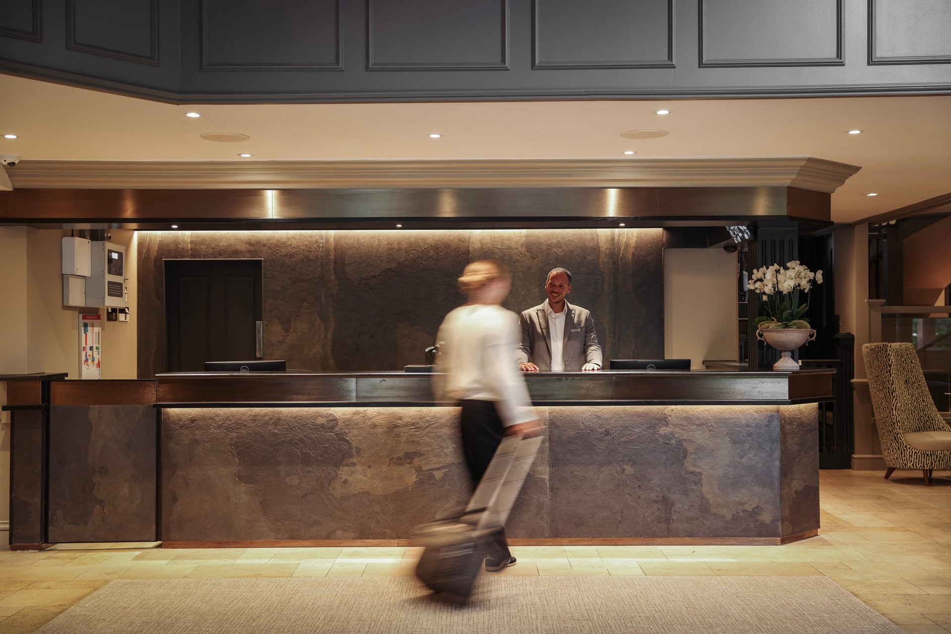 Modern hotel lobby with a blurred guest carrying luggage approaching a dark wood reception desk staffed by two clerks, with recessed lighting and warm tones.