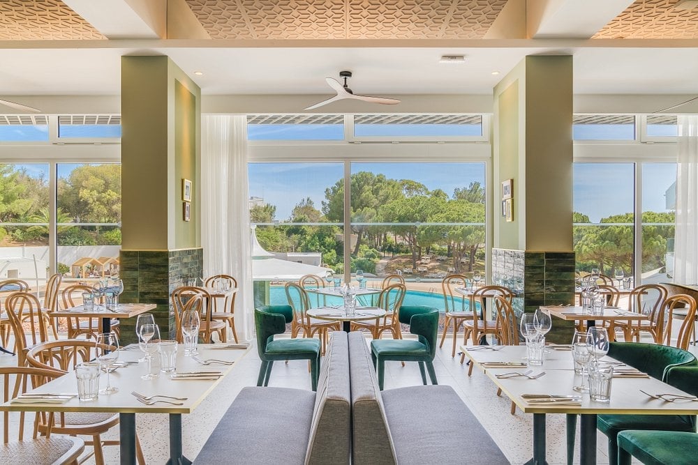 Bright modern restaurant dining room with natural wood chairs, white tablecloths, and large windows overlooking landscaped grounds and trees