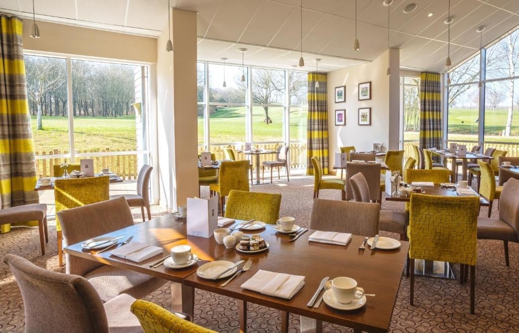 Bright dining room with large windows overlooking green landscape, wooden tables, mustard and beige chairs, place settings, and natural lighting